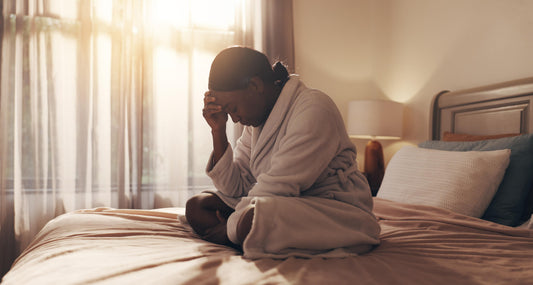 Black woman sitting on a bed in soft morning light, holding her head in her hand with a tired and overwhelmed expression, reflecting emotional exhaustion, burnout, and mental fatigue.