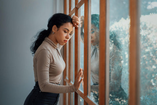 Pensive woman standing by a window with her head lowered and hand against the glass, reflecting emotional exhaustion, grief, or quiet overwhelm.