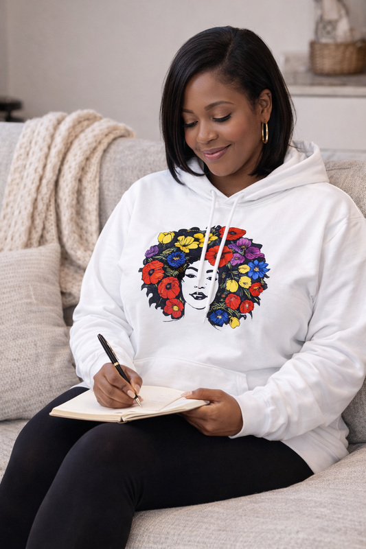Black woman  wearing a white floral portrait hoodie while journaling on a cozy beige couch in a softly lit living room