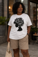 Plus size Black woman wearing a white afrocentric floral silhouette t-shirt in modern city setting.